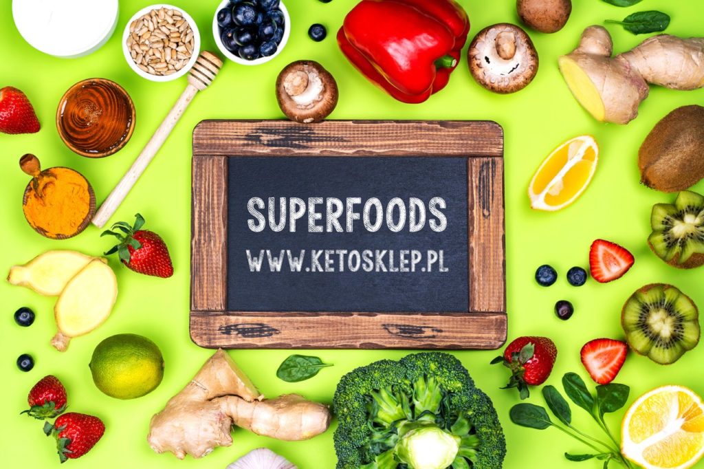 Tablica z napisem SUPERFOODS na środku na zielonym tle. W tle porozrzucane różne produkty możliwe do zjedzenia na diecie keto.